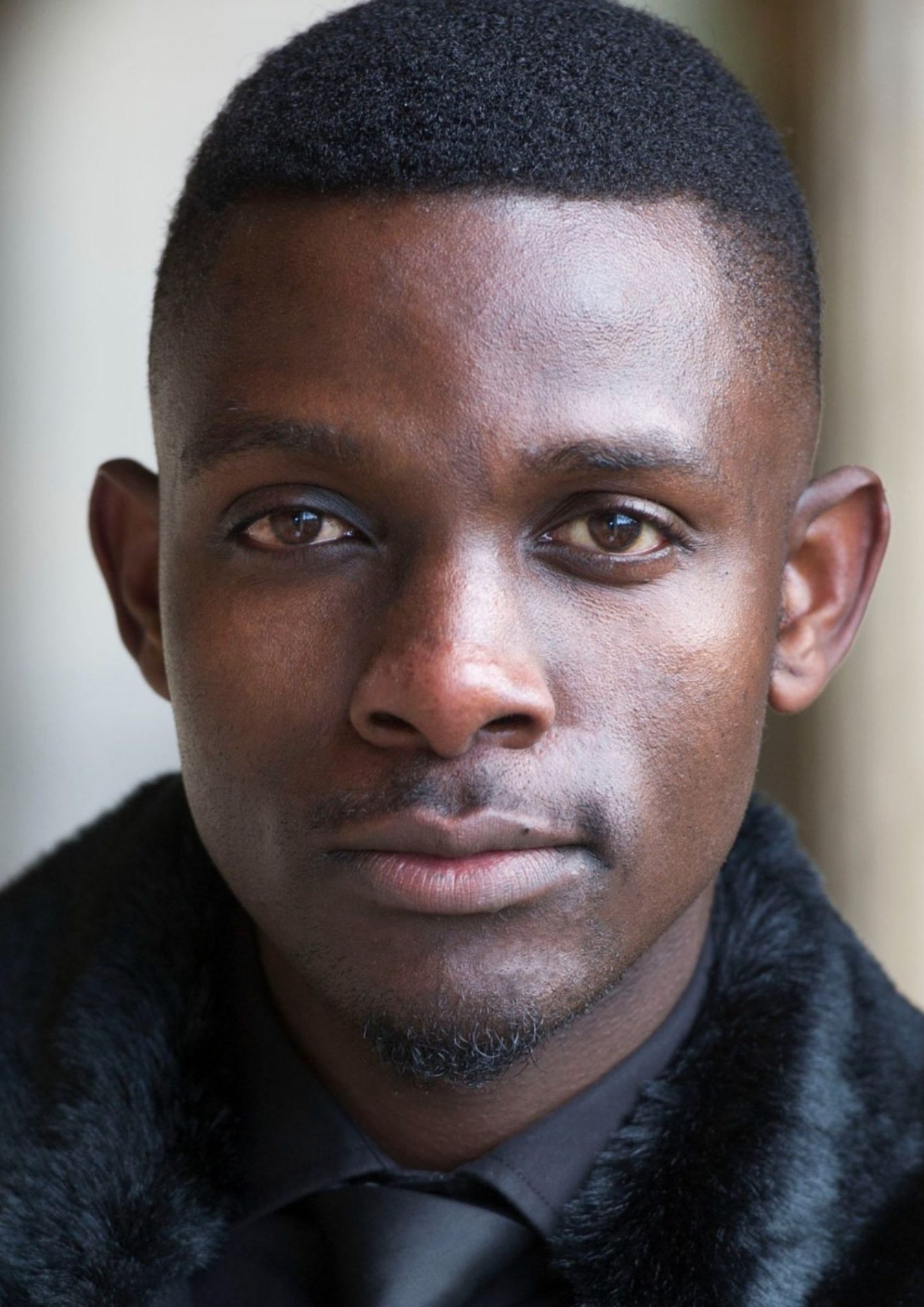 An actor with short black hair has a neutral facial expression. They are wearing a black shirt and black tie with a black fur coat.