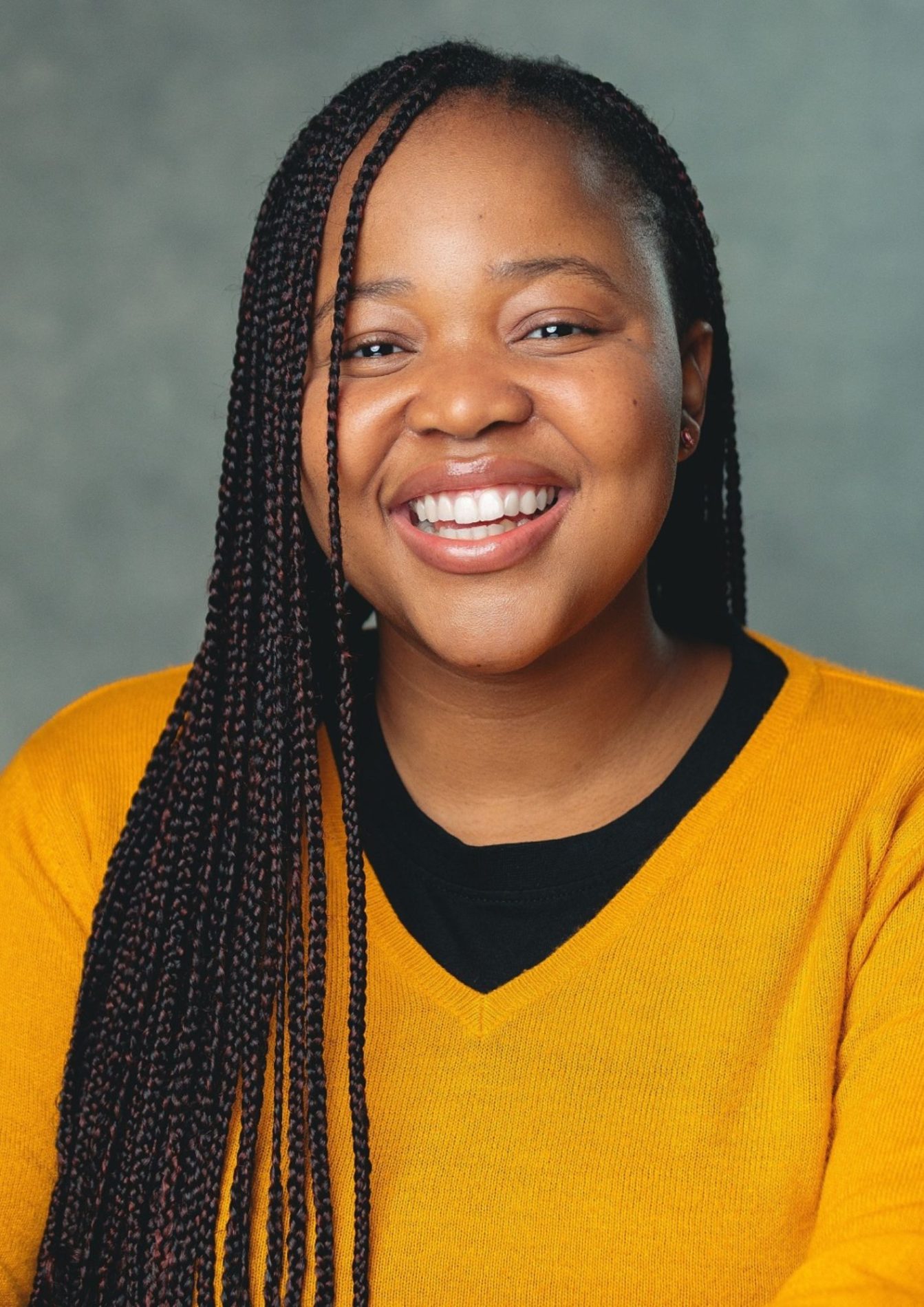 An actor with long black hair in braids has a happy facial expression and is smiling. They are wearing an orange sweater with a black shirt underneath.