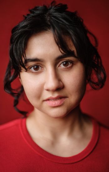 A headshot of an actor with short black hair and a red shirt is wearing a serious expression.