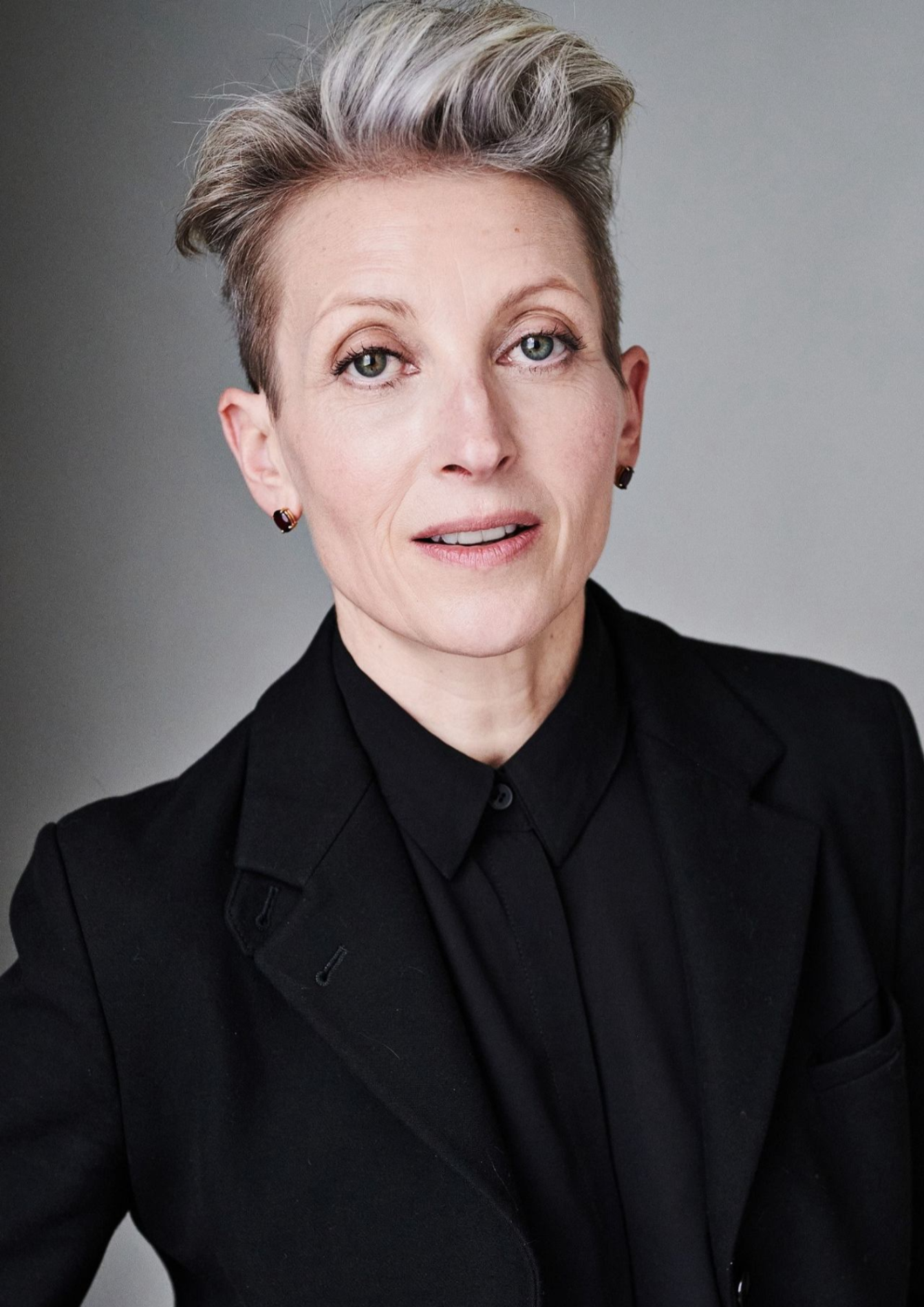 An actor with short grey and brown hair has a neutral facial expression. They are wearing a black button down top.