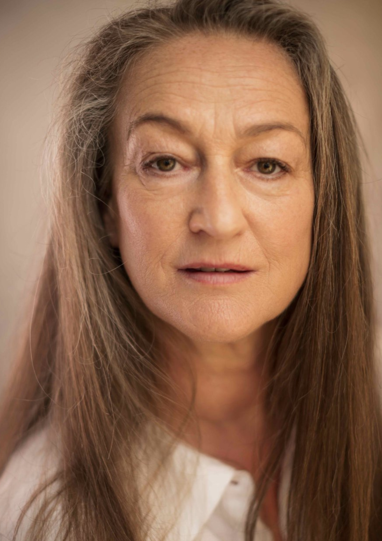 An actor with long brown hair has a neutral facial expression. They are wearing a white collared shirt.