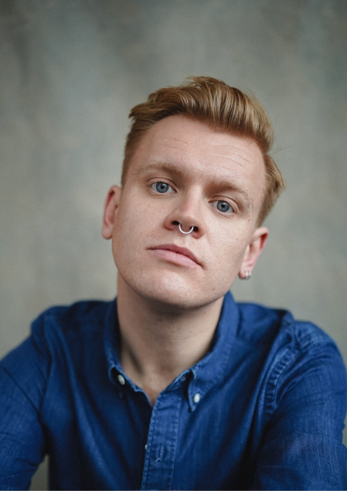 An actor with short strawberry blonde hair and a nose ring has a neutral facial expression. They are wearing a denim button down shirt.