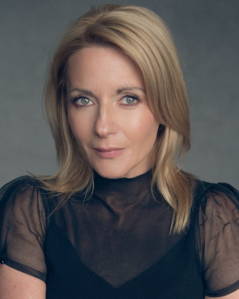 An actor with medium length blonde hair has a neutral facial expression. They are wearing a black mesh top.