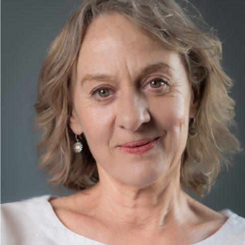 An actor with shoulder length blonde hair has a pleasant facial expression with a slight smile. They are wearing a white top.