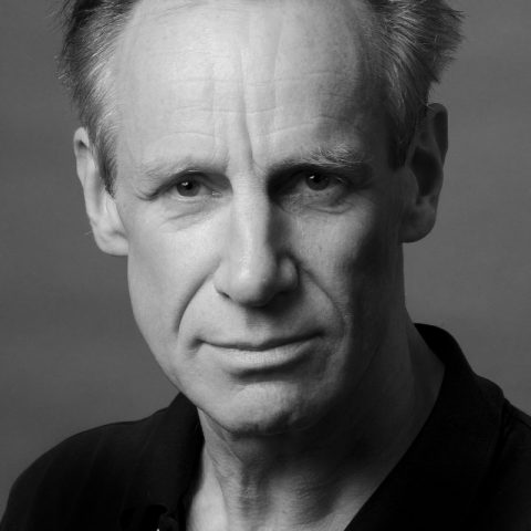 An actor with short hair is in greyscale. They have an intense facial expression and are wearing a dark top.