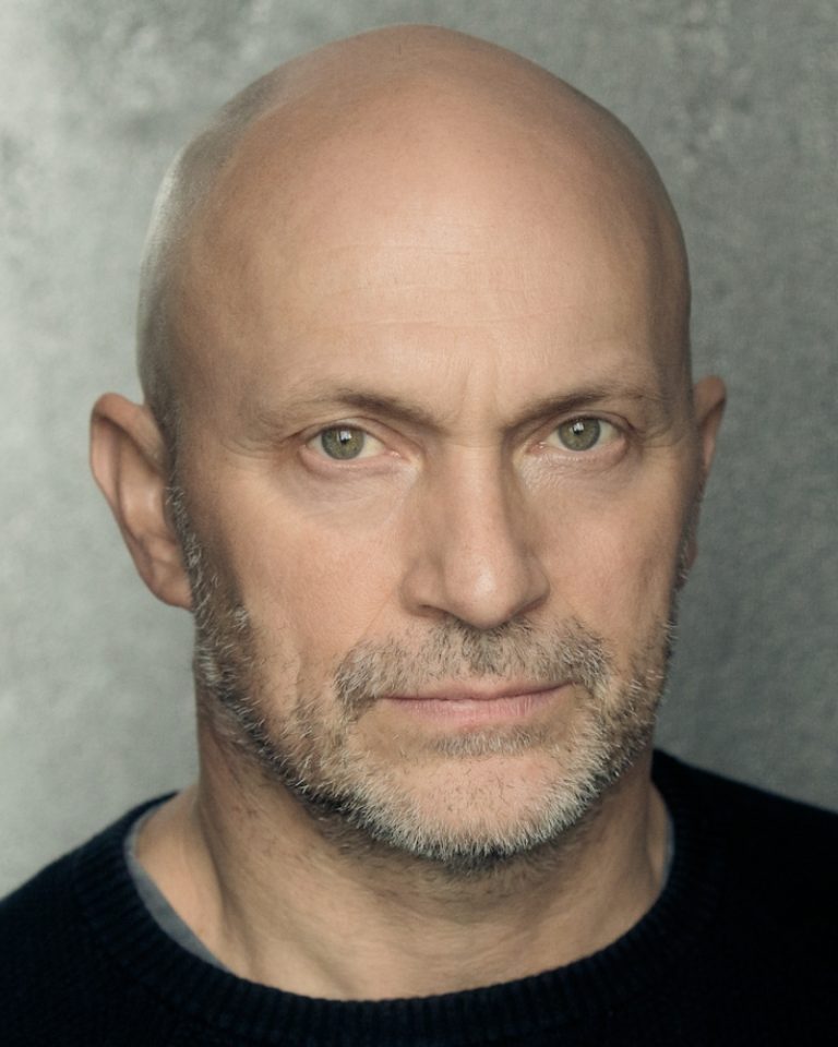 An actor who is bald and has a beard has an intense facial expression. They are wearing a black top.