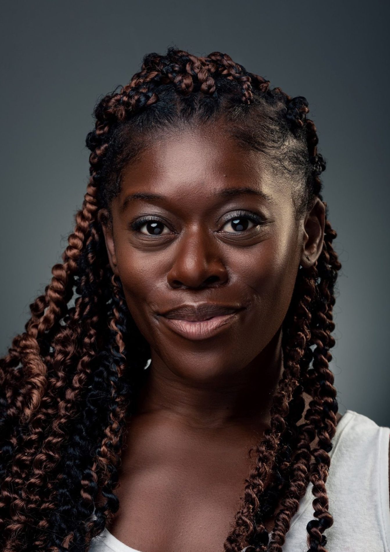 A headshot of an actor with long black hair with brown highlights. They are wearing a light grey shirt. They are wearing a happy expression.