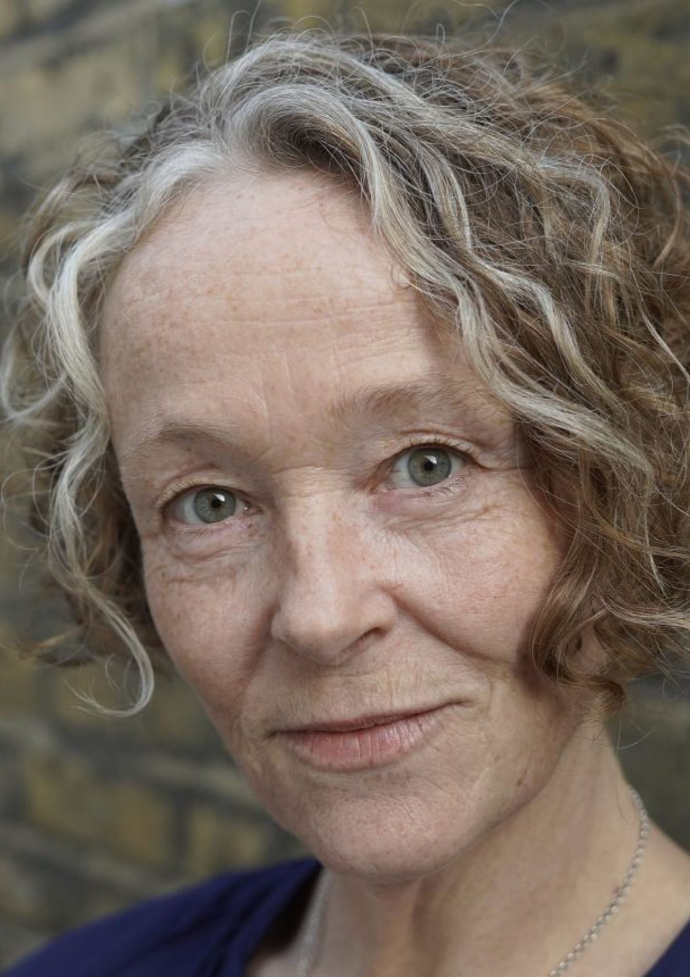 A headshot of an actor with brown and grey medium-length hair. They are wearing a black shirt and a silver necklace. They have a slight smile on their face.