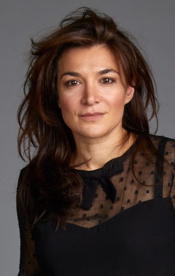 A headshot of an actor with medium-length dark brown hair. They are wearing a black blouse and have a neutral expression on their face.