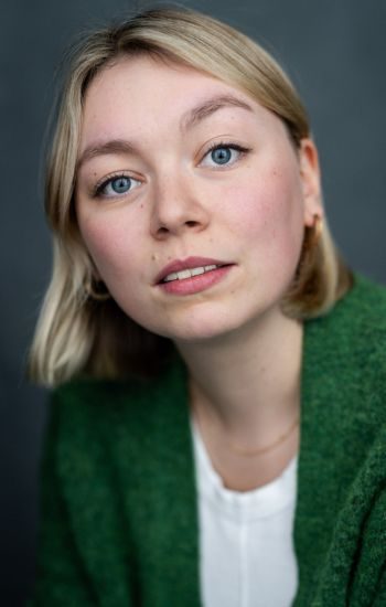 A headshot of an actor with medium blond hair and blue eyes. \they are wearing a white shirt and dark green sweater. They are wearing a blank expression.