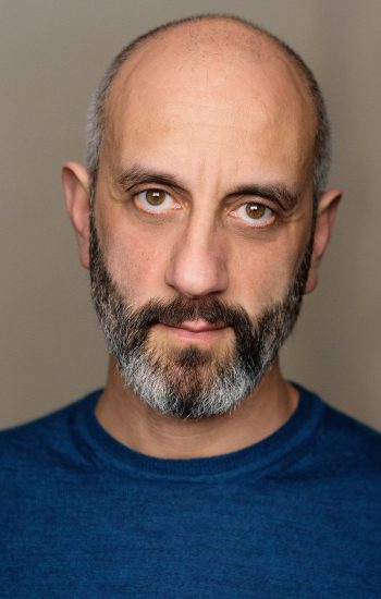 A headshot of an actor with a black and grey beard and blue shirt. He is wearing a mischievous expression.